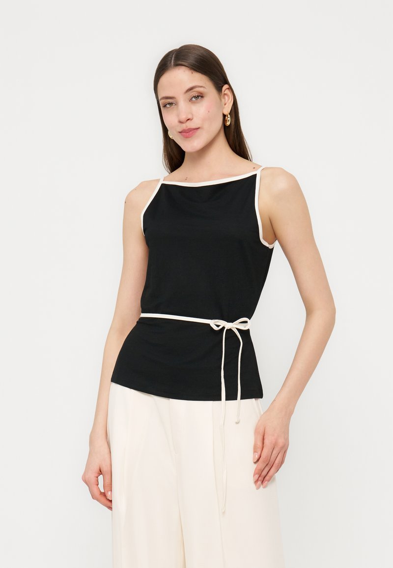 Γυναίκα που φοράει μαύρο αμάνικο τοπ με λευκή περίμετρο και ζώνη με κορδόνι, συνδυασμένο με λευκό παντελόνι φαρδιάς γραμμής, στέκεται μπροστά σε ένα μονόχρωμο φόντο.