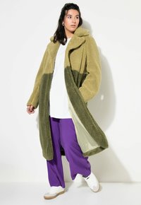 Cappotto lungo bicolore realizzato in materiale morbido e peloso in verde oliva e salvia. Presenta un cappuccio e maniche ampie. Indossato aperto sopra un top bianco e pantaloni viola.
