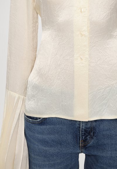Bluză de culoare crem, cu o textură plisată, prevăzută cu o deschidere centrală cu nasturi și mâneci lungi plisate, asortată cu blugi din denim albastru.
