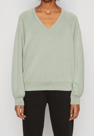 Osoba ubrana w jasnozielony, długi sweter z dekoltem w serek i elastycznymi mankietami, oraz czarne spodnie, widoczna od ramion do bioder.