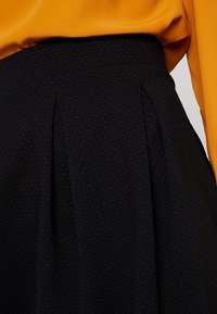 Jupe noire texturée taille haute avec des plis, portée avec un haut orange vif rentré à l'intérieur.