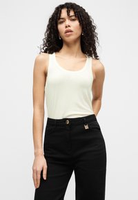 Ανοιχτό πράσινο τοπ με λαιμόκοψη scoop, σε συνδυασμό με μαύρο παντελόνι που διαθέτει χρυσά εξαρτήματα και ορατές τσέπες. Το μοντέλο έχει σγουρά μαλλιά.