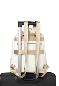 White textured backpack with beige straps, featuring a front zipper pocket, resting on a black suitcase handle. Minimalist design.
