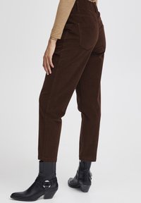 Osoba ubrana w spodnie z wysokim stanem w ciemno-brązowym kolorze i czarne botki za kostkę, stoi z jedną ręką swobodnie opuszczoną przy boku.