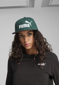 Căciulă verde tip snapback cu logo alb "PUMA" și grafică a pisicii; vizor plat; purtată cu o cămașă neagră cu un mic logo pe piept.