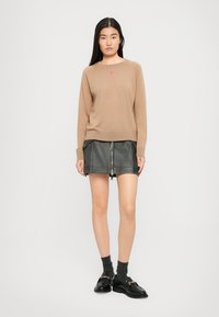 Kvinne som har på seg en tan-genser med et lite rosa hjerte, svart skinn mini-skjørt med frontglidelås, mørke sokker og svarte loafers mot hvit bakgrunn.