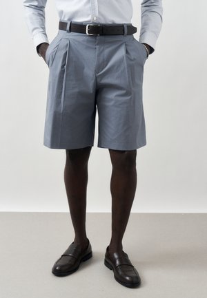 Mężczyzna ubrany w jasnoniebieskie, dopasowane szorty, czarny pasek, jasnoniebieską koszulę, czarne mokasyny, stoi z rękami w kieszeniach.