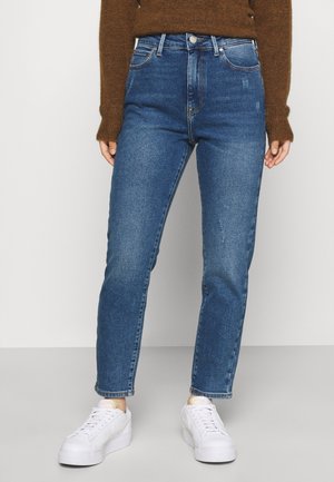 Person iført blå højtaljede, slim-fit jeans, brun sweater og hvide sneakers, stående mod en ensfarvet lys baggrund.
