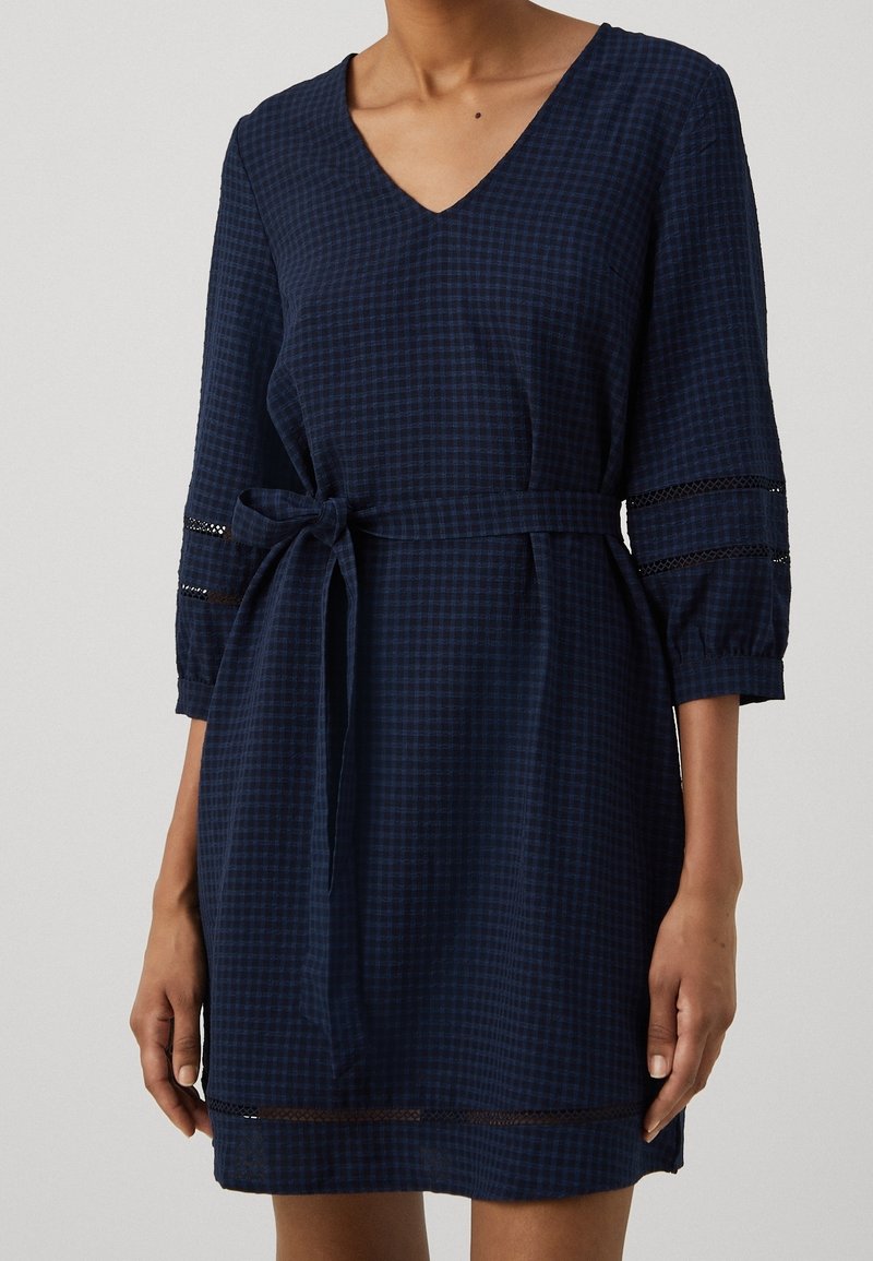 Robe bleu marine longueur genoux avec petit motif à carreaux, col en V, manches trois-quarts, ceinture à nouer à la taille et insertions en dentelle sur les manches et l'ourlet.