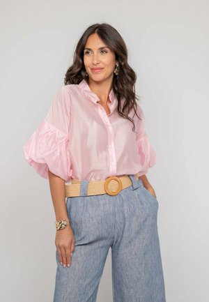 Femme aux cheveux ondulés foncés portant un chemisier rose clair à volants, un pantalon bleu taille haute, une ceinture beige à boucle ronde et un bracelet en or.