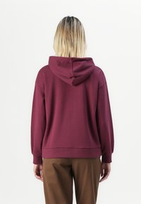 Burgundy hoodie med en rymlig huva, ribbade ärmslut och nederkant. Mjukt material med en avslappnad passform, kombinerad med bruna byxor.