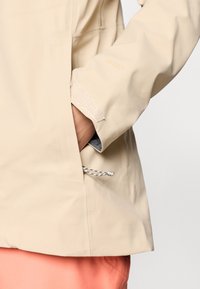 Μπεζ αδιάβροχο μπουφάν με υφασμάτινη υφή, που διαθέτει πλαϊνές τσέπες με κορδόνι και ρυθμιζόμενους καρπούς με βέλκρο.