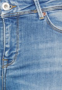 Tecido de denim azul claro com uma textura desbotada, costura dourada e um botão prateado na cintura. Apresenta uma costura de bolso lateral.