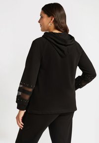 Svart hoodie med rundad halsringning, ärmar i nät med små nitar, och en huva med dragsko. Mjuk tygtextur.