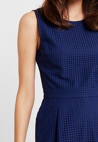 Bezramenné námořnické modré šaty s texturovaným mřížkovaným vzorem, které má na sobě osoba s hnědými vlasy, zachycena od ramene po pas.