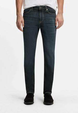 Mężczyzna ubrany w ciemnoniebieskie jeansy slim fit i czarne buty, stojący z rękami swobodnie opuszczonymi wzdłuż ciała na białym tle.