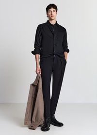 Schwarzes Hemd mit Knopfleiste und Tasche, kombiniert mit schwarzer Hose. Ein beiger Mantel wird gehalten, und glänzende schwarze Stiefeletten werden getragen.