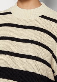 Pull rayé crème et noir, doté d'un col rond côtelé et d'une texture tricotée ample avec de larges rayures horizontales.