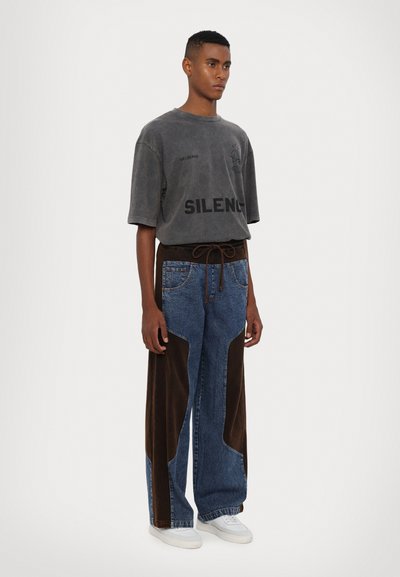 T-shirt gris surdimensionné avec "SILENC" imprimé, associé à un pantalon en denim bleu avec des panneaux en velours côtelé marron sur les côtés et une taille à cordon.