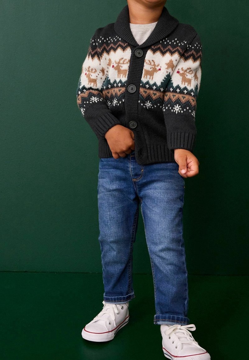 Enfant portant un pull noir boutonné avec des motifs de rennes, un jean bleu et des baskets blanches, debout sur un fond vert foncé.