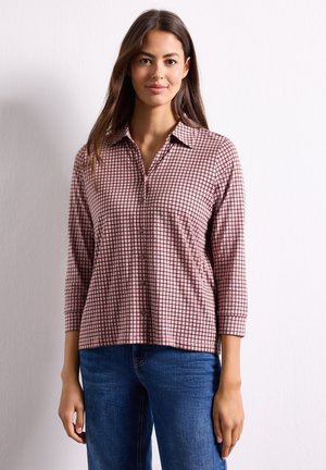 Femme aux longs cheveux bruns portant une chemise à carreaux rose et noire boutonnée et un jean bleu, debout devant un fond blanc.