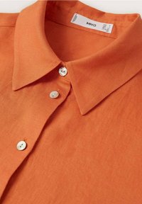 Camisa de lino naranja de manga corta con un cuello clásico, que presenta botones blancos y una tela texturizada. Etiqueta de la marca visible dentro del cuello.