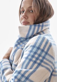 Fleece-takki, jossa on sinivalkoinen ruudullinen kuvio, korkea kaulus ja teksturoitu kangas, roikkumassa hartioilla.