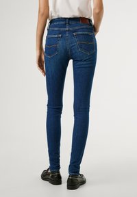Pantalones vaqueros skinny azul oscuro con una textura suave, que presentan dos bolsillos traseros y detalles de costura sutiles. Combinados con zapatos negros.