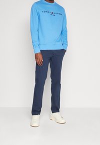 Felpa azzurro chiaro con la scritta "TOMMY HILFIGER", abbinata a pantaloni blu navy e sneakers bianche. La felpa presenta un colletto a girocollo e polsini a costine.