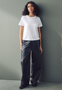 Wit katoenen T-shirt met een getextureerde kraag, gecombineerd met hoge taille zwarte leren wijde broekspijpen en zwarte schoenen. Effen achtergrond.