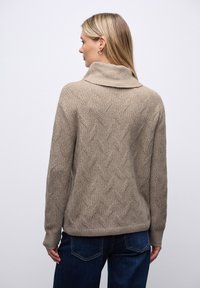 Beżowy sweter z dzianiny z wysokim kołnierzem, ozdobiony teksturowanym wzorem w zygzak oraz ściągaczem na dole, w połączeniu z ciemnoniebieskimi dżinsami.