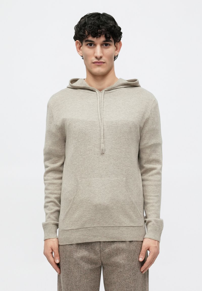 Beige hættetrøje med blød tekstur, front kangaroo lomme, lange ærmer, snøre lukning, parret med strukturerede brune bukser.