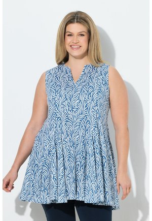 Uśmiechnięta kobieta o blond włosach do ramion, ubrana w bezrękawnikową tunikę w niebiesko-biały wzór, na ciemnych spodniach, stojąca na tle białej ściany.