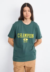 Koszulka bawełniana w leśnozielonym kolorze z wyrazistym żółtym napisem i logiem "CHAMPION", z okrągłym dekoltem i krótkimi rękawami.