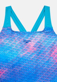 Μαγιό με ζιγκ-ζαγκ μοτίβο σε αποχρώσεις του μπλε και του ροζ. Οι τιράντες είναι ανοιχτό μπλε και σταυρωτές στο πίσω μέρος. Το λογότυπο της μάρκας είναι εμφανές.