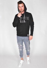 Svart hoodie med "TOP GUN" grafik, ribbat avslut och dragsnören. Bärs med ljusblå slitna jeans och vita sneakers.