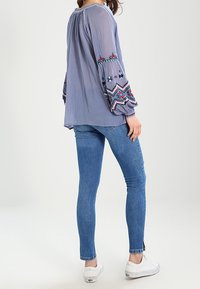Blusa de manga larga azul claro con patrones bordados en las mangas, combinada con jeans ajustados azules y zapatillas blancas.