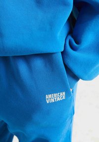 Spilgti zili sporta bikses ar roku kabatā un uzrakstu "AMERICAN VINTAGE" baltā krāsā netālu no kabatas.