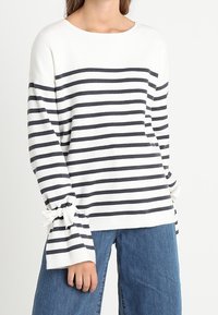 Donna che indossa una maglietta bianca a maniche lunghe con strisce orizzontali nere e dettagli a fiocco sui polsini a campana, abbinata a jeans in denim blu.