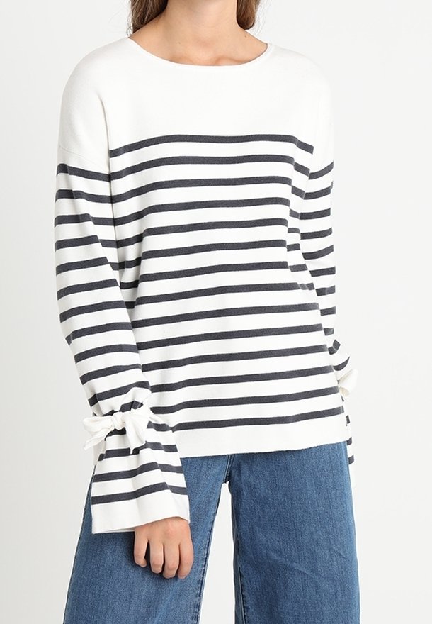 Donna che indossa una maglietta bianca a maniche lunghe con strisce orizzontali nere e dettagli a fiocco sui polsini a campana, abbinata a jeans in denim blu.