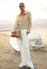 Braun-weiße Hahnentrittmuster-Jacke, weiße weit geschnittene Hose, hält eine beige gesteppte Tasche, mit reflektierenden Oberflächen im Hintergrund.