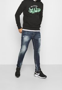 Sudadera negra con logo verde, camiseta blanca debajo, jeans azules desgastados y zapatillas negras con detalles blancos. Modelo de pie contra un fondo blanco.