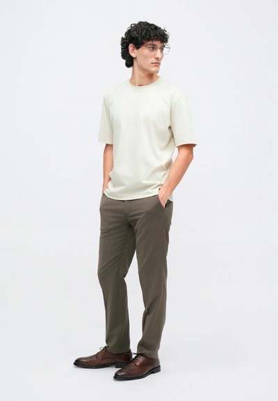 Beige rövid ujjú póló olíva zöld nadrággal és barna bőr cipővel párosítva. Az öltözet laza szabású és sima textúrájú.