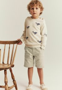 Striktrøje i kremfarvet med blåt hvalmønster, rund halsudskæring og knapdetaljer på skulderen. Lys beige shorts og hvide sko.