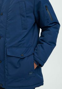 Navy blue padded jacket featuring a front button closure, multiple pockets, textured fabric, and a zippered sleeve pocket detail.