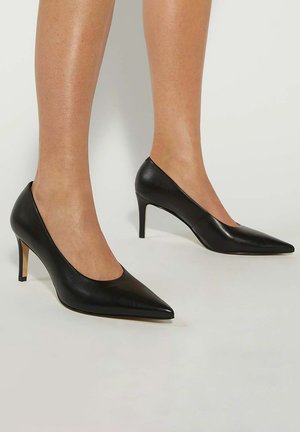 Benen in zwarte leren puntige hakken met slanke stiletto-hakken op een lichte achtergrond.