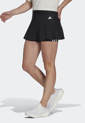 Femme portant un skort noir Adidas et des baskets blanches, debout avec une main sur la hanche et un pied légèrement levé.