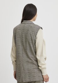 Gilet à carreaux gris et noir avec une manche en maille crème. Présente un col ajusté et un design dos ouvert. Tissu texturé.