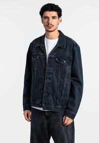 Giacca di jeans scura con una vestibilità comoda, chiusura a bottoni, tasche sul petto e cuciture a contrasto, indossata sopra una maglietta bianca. Design semplice.