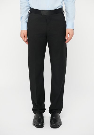 Muž oblečený v černých společenských kalhotách s páskem na nastavení velikosti, světle modré košili a černých kožených botách, stojící vzpřímeně na bílém pozadí.